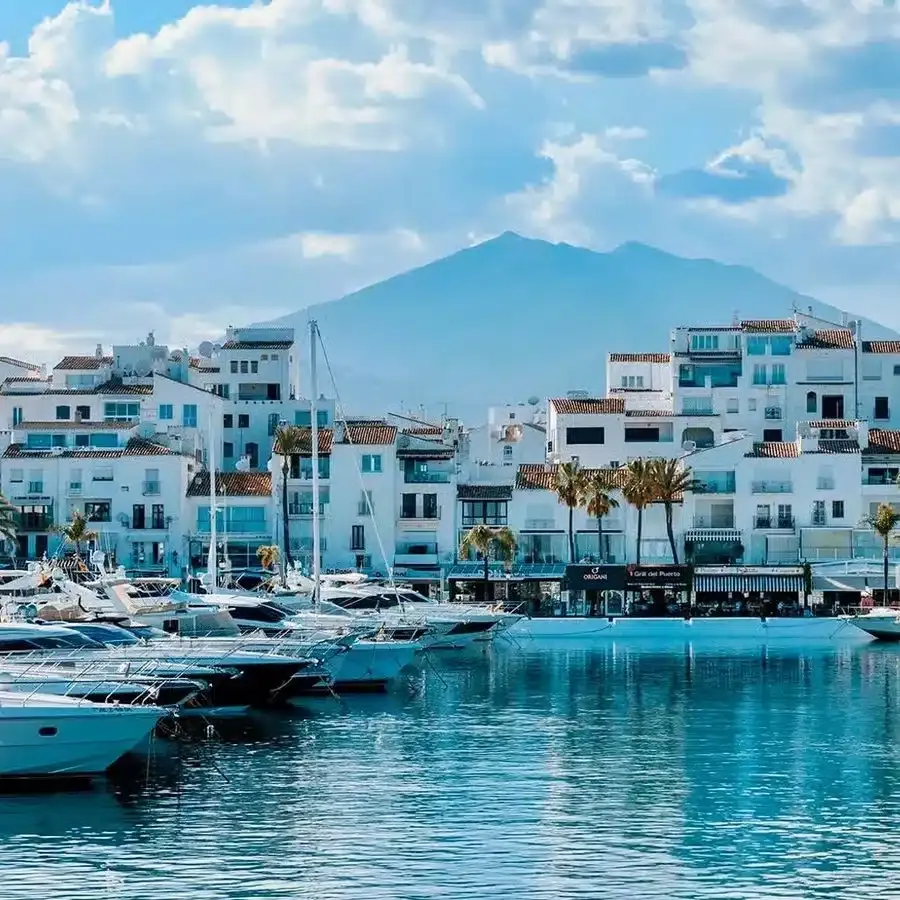 Luxury yachts in Puerto Banús marina with white buildings and a mountain backdrop under a blue sky, illustrating the high-end appeal for real estate investment in Spain.
